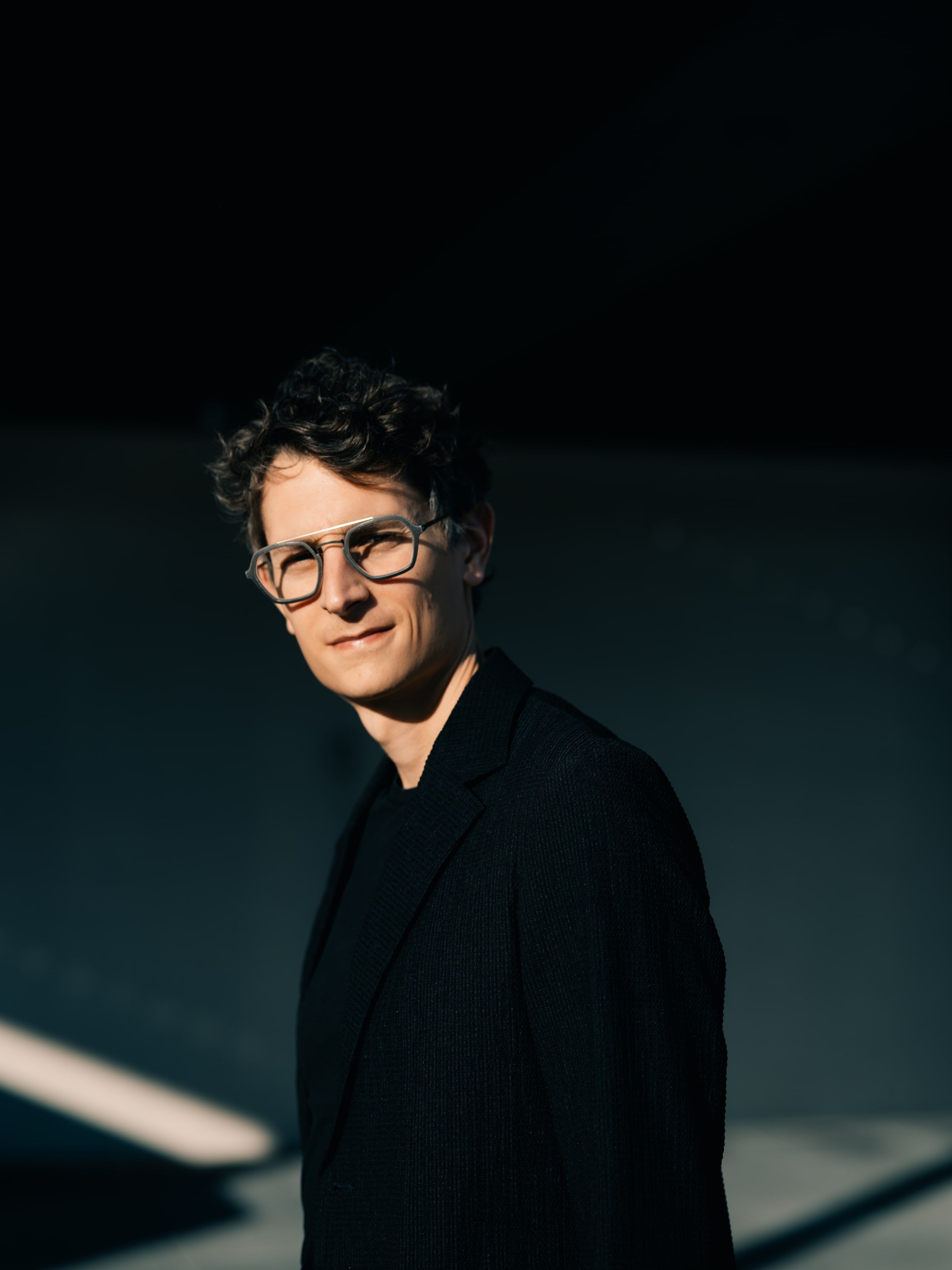 Matthew Claudel in a black jacket, half lit by sunlight against a dark wall, looking at the camera.